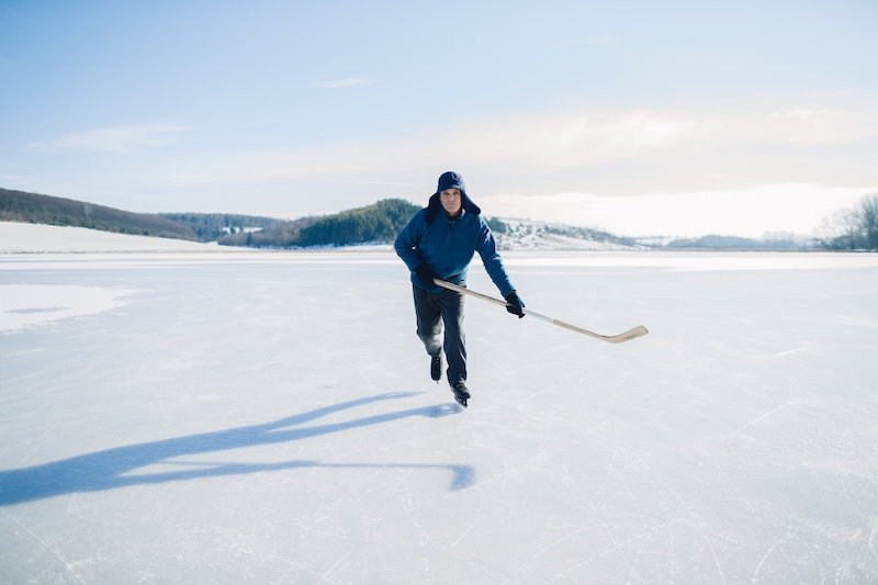 Senior man on ice skates with hockey stick on frozen lake in winter. Hobby concept of elderly people.